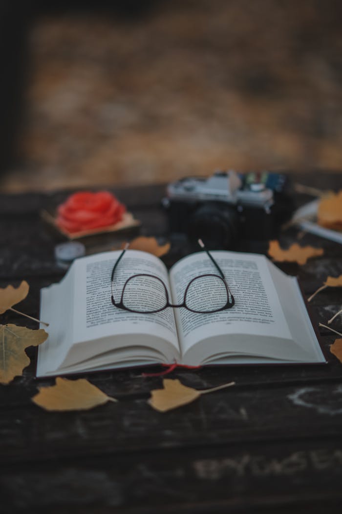 Open book with glasses on a wooden table surrounded by autumn leaves and vintage camera.